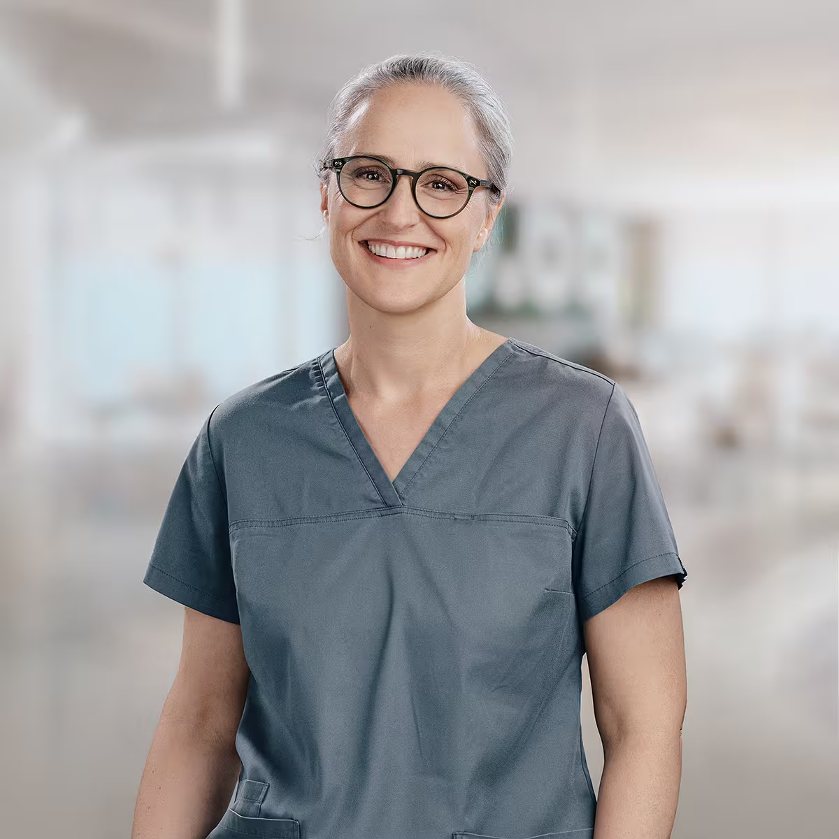 Janine Suntz mit grauem Haar und Brille trägt graue medizinische Arbeitskleidung.